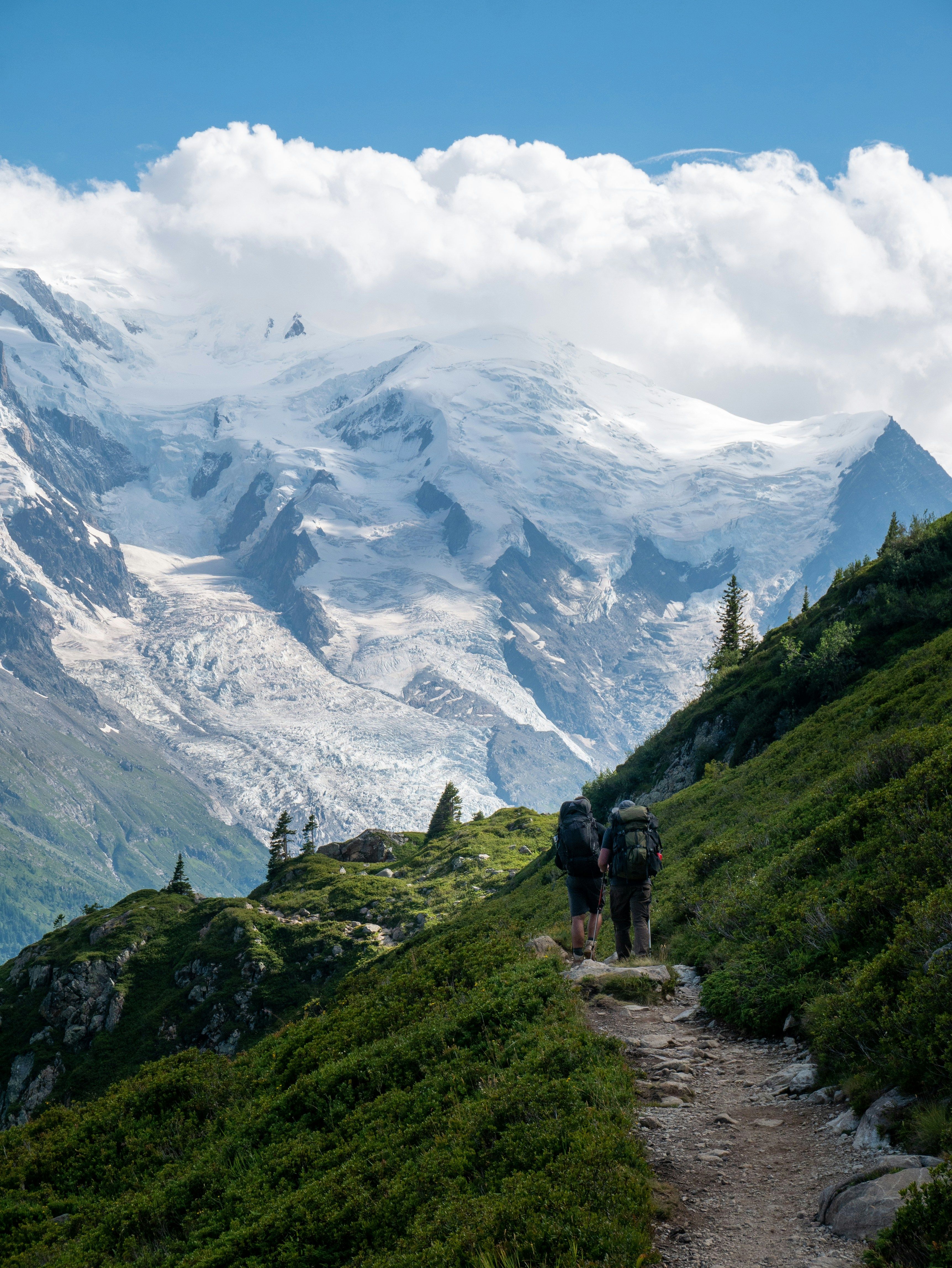 Two hikers on a mountain trail facing a snow-capped peak